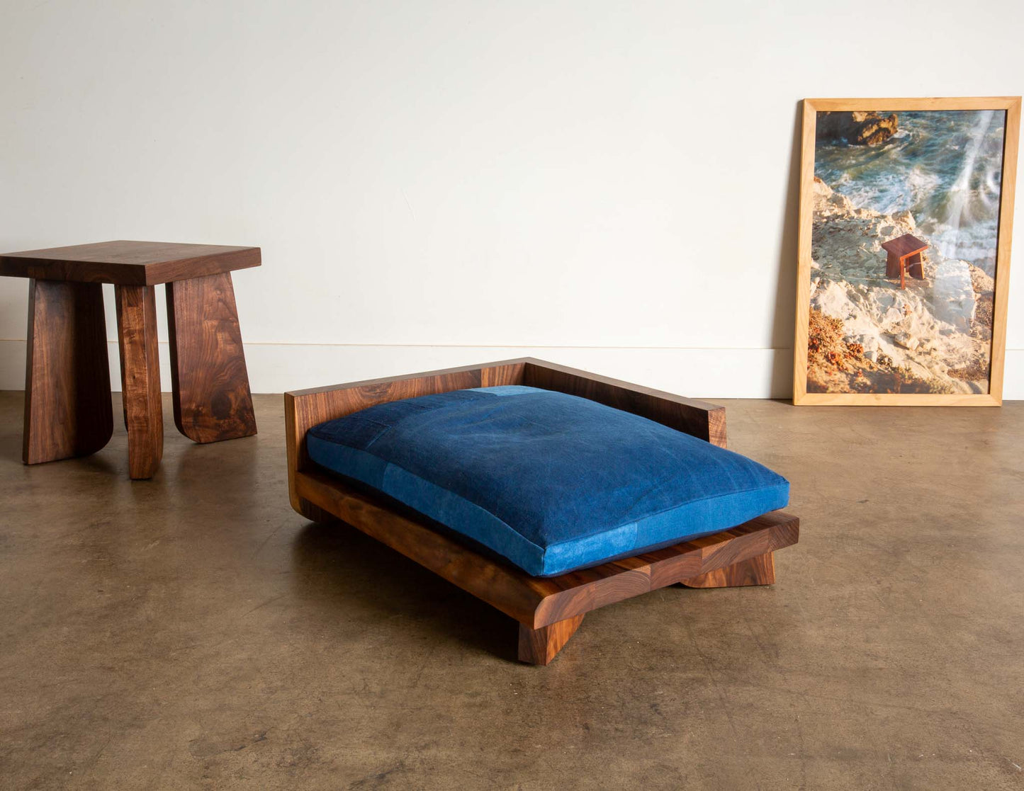 A blue indigo cushioned dog bed with a walnut brown frame, placed on a concrete floor against a wall with a framed picture and a smaller picture frame, with a Walnut Temple Stool visible in the background.