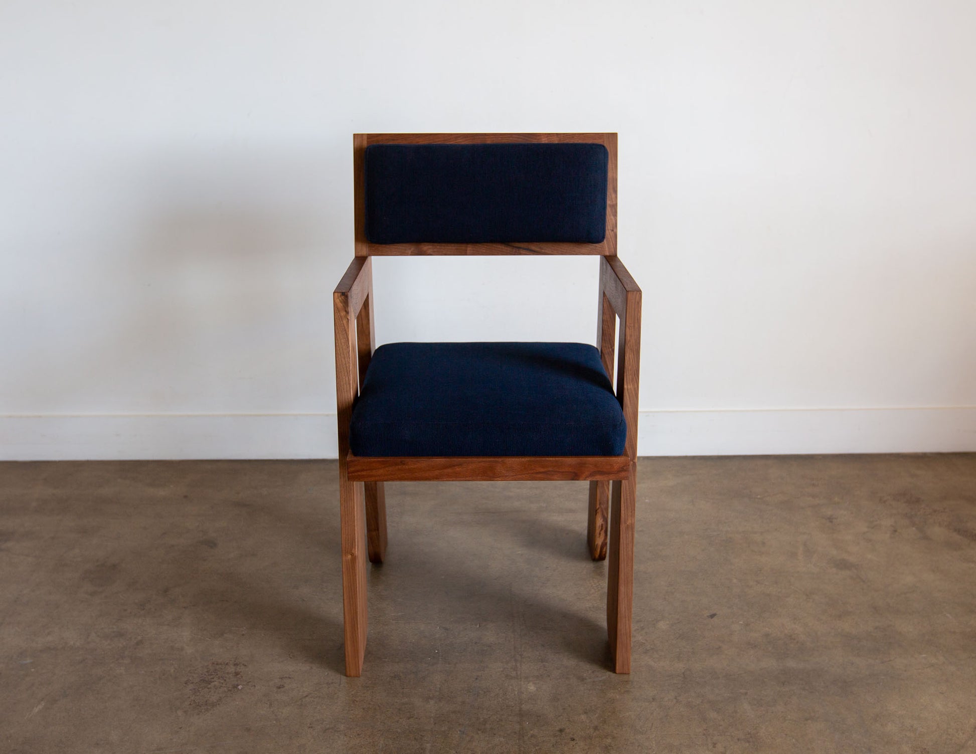 A front view of the hand-built Snyder DePass Walnut Temple Upholstered Chair, featuring a minimalist design with a rich, dark fabric and a seamless solid walnut frame.