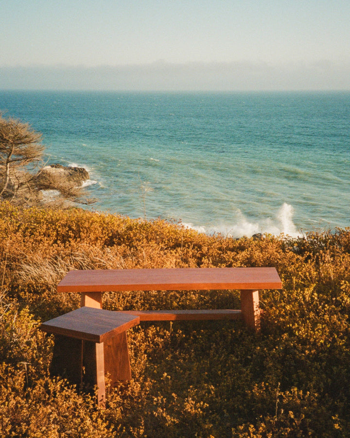 The Snyder DePass Temple Bench and Stool, both crafted from solid Walnut, are shown together on a sandy Malibu beach, highlighting their minimalist design and natural materials.