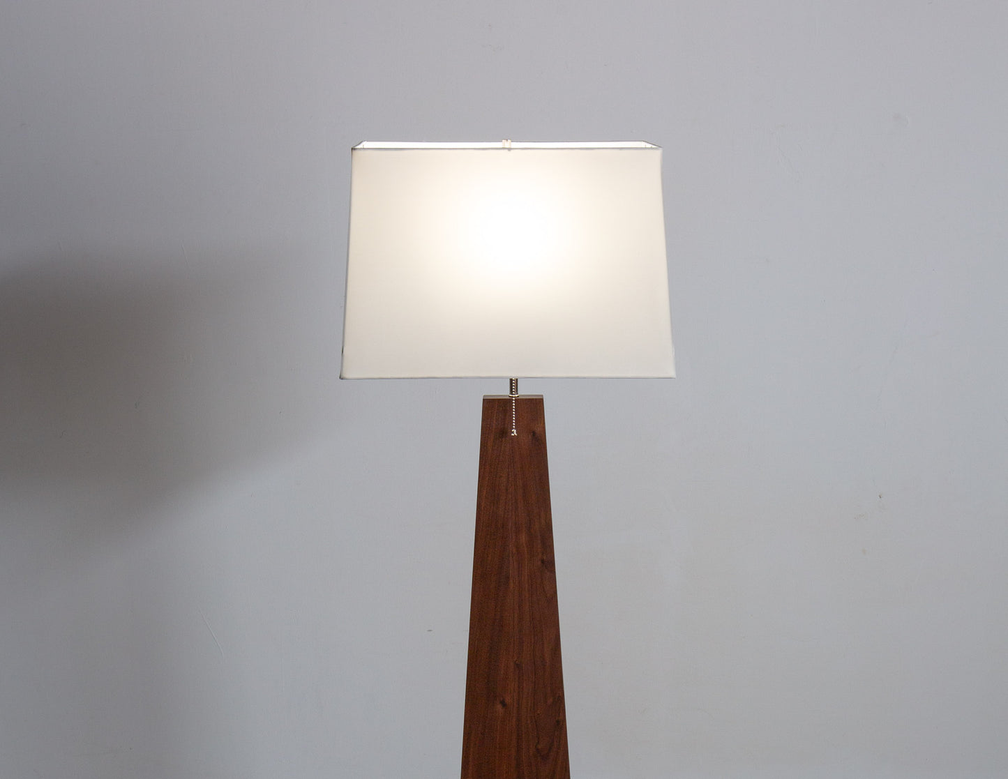 A close-up of the Pas Floor Lamp, featuring a tall, tapered wooden base and a white rectangular shade. The lamp is on, and a pull chain hangs from the socket.