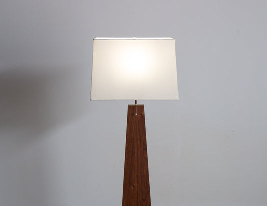 A close-up of the Pas Floor Lamp, featuring a tall, tapered wooden base and a white rectangular shade. The lamp is on, and a pull chain hangs from the socket.